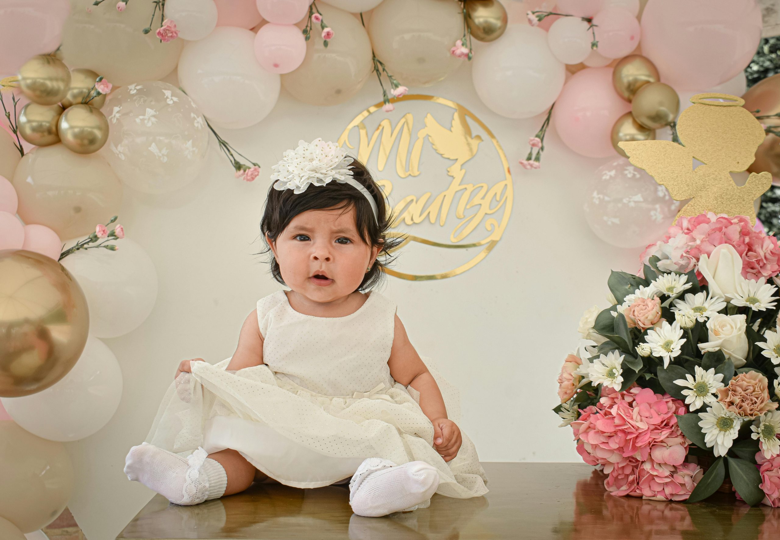 Adorable baby in a white dress celebrating with festive balloons and flowers in Colombia.